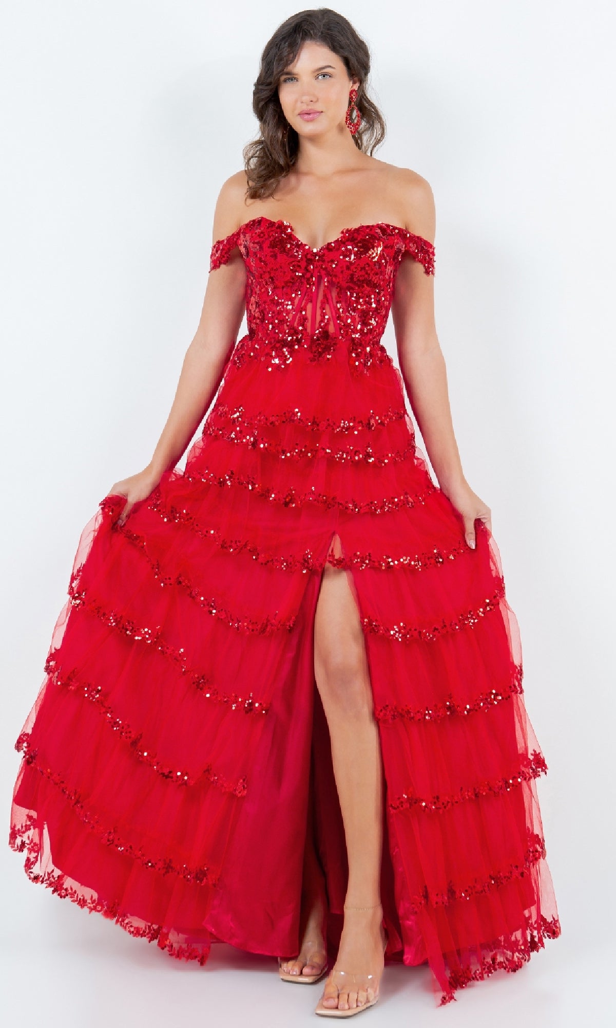A woman stands against a plain white background wearing the Ruffle Prom Ball Gown: Cinderella Couture 8228J—an off-the-shoulder, floor-length red dress with layered ruffles, sequin embroidery, and a front slit.
